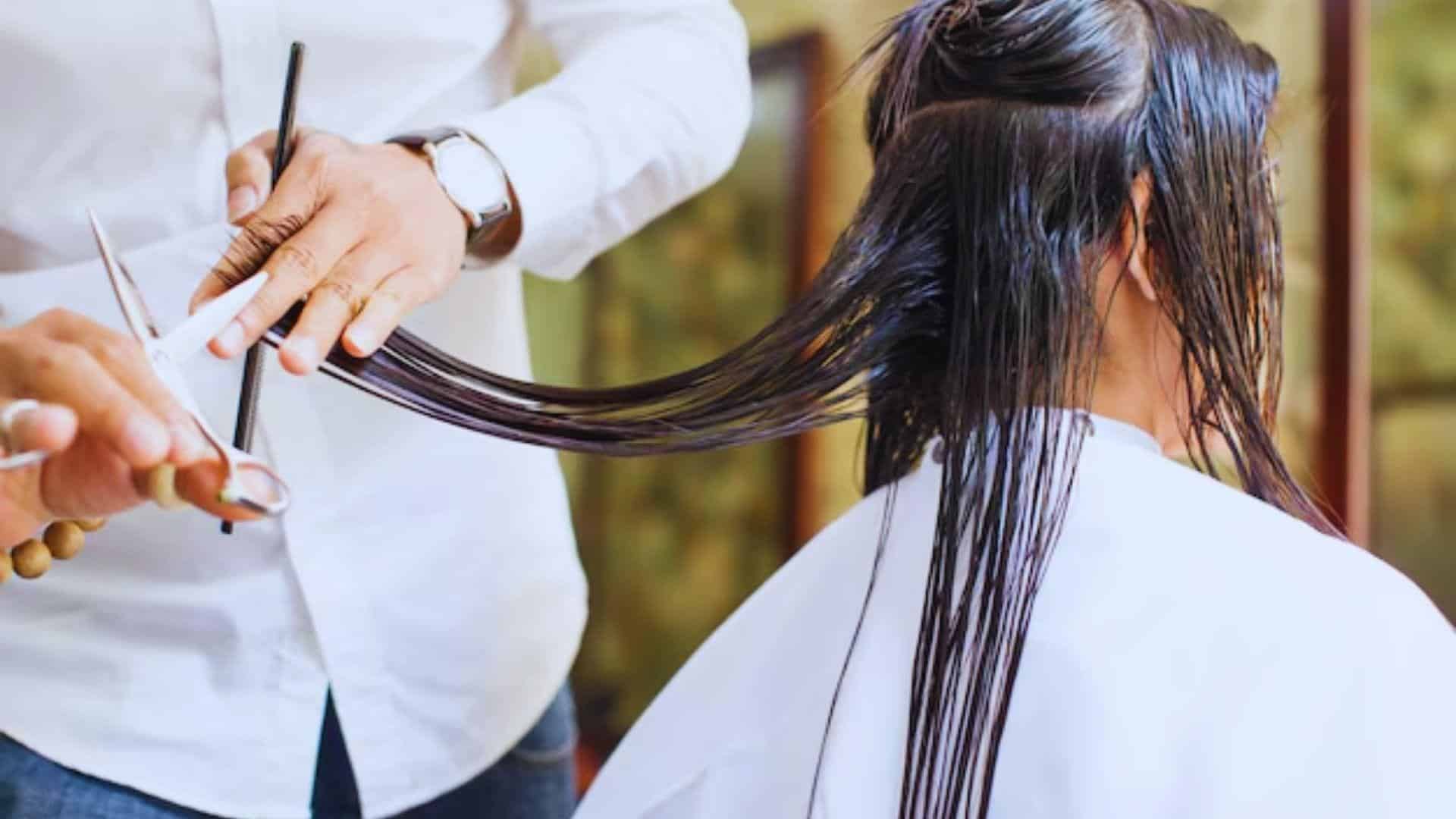 A woman sits in a salon chair as a man styles her hair with scissors.