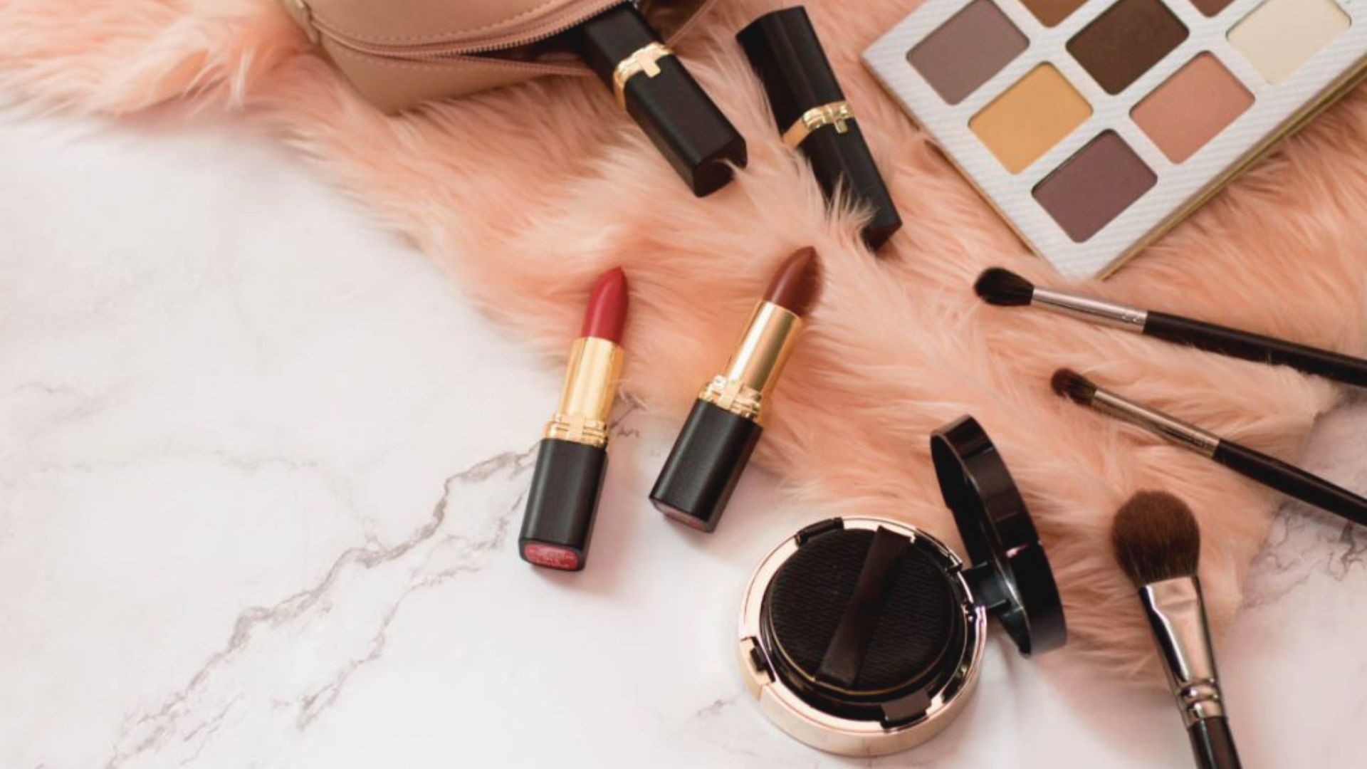 A pink fur coat, makeup brushes, and a purse are artistically arranged on a white marble surface, highlighting fashion elements.