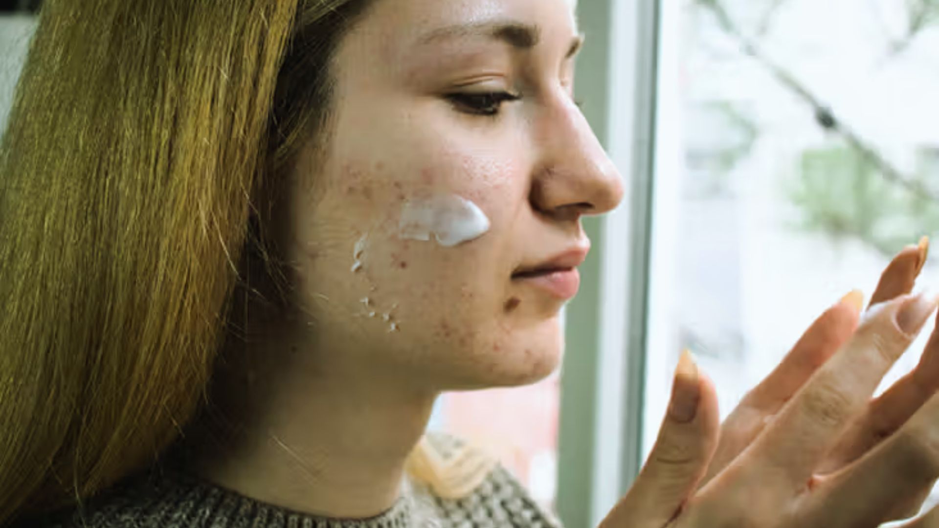 A woman gently applying cream to her face, focusing on skincare in a well-lit bathroom setting.