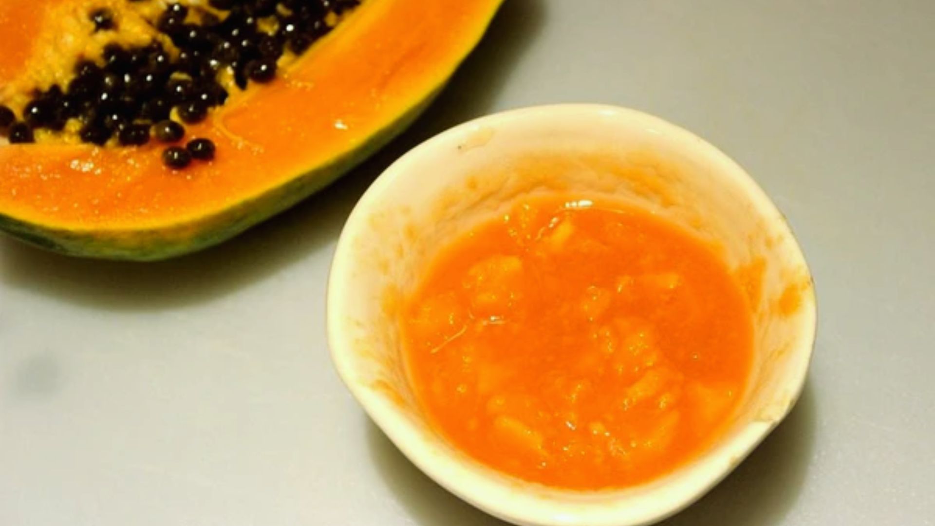 A bowl of vibrant orange sauce placed beside a ripe papaya on a neutral surface.