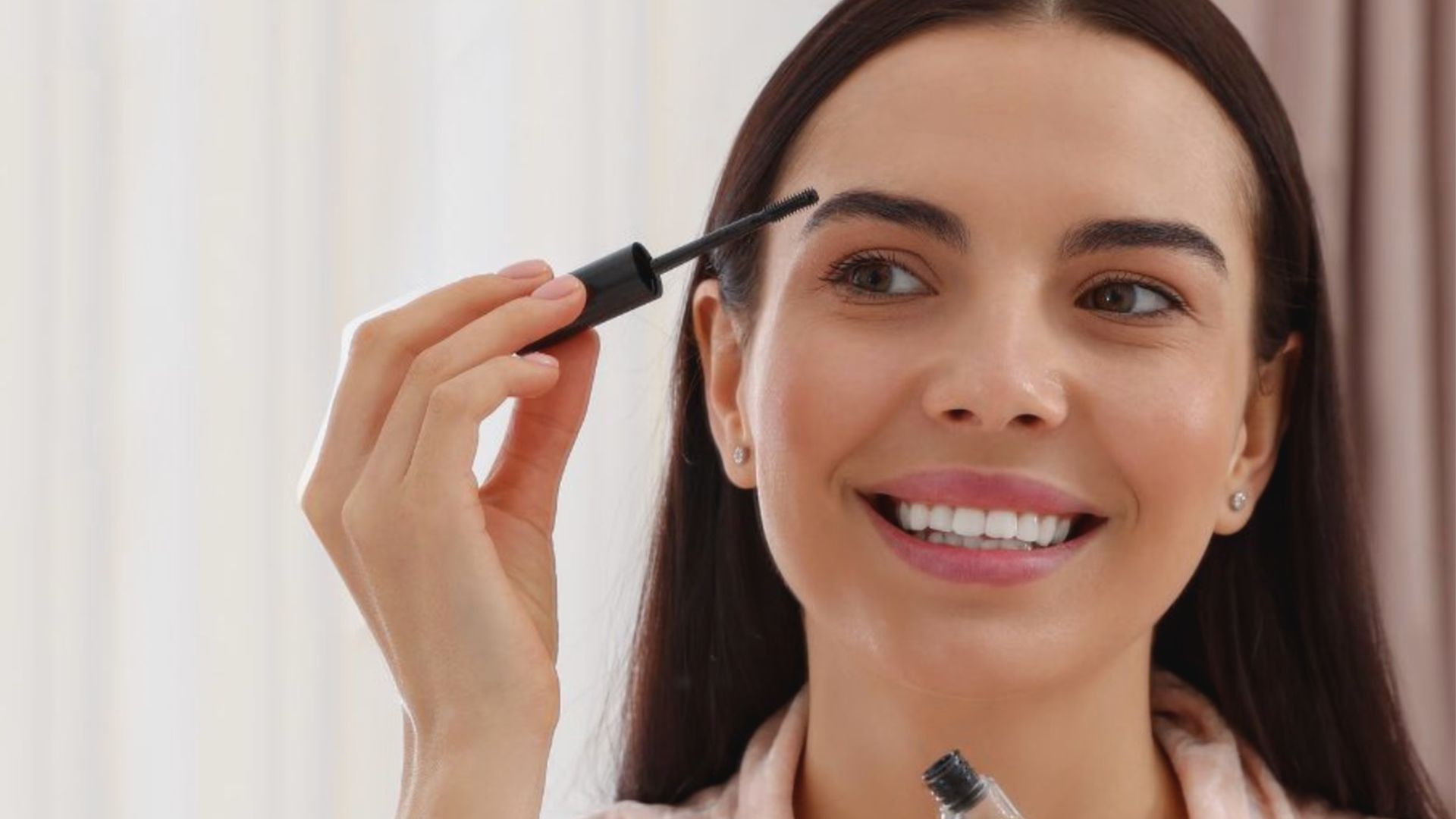 A woman joyfully applies mascara with a brush, showcasing her beauty routine in a cheerful atmosphere.