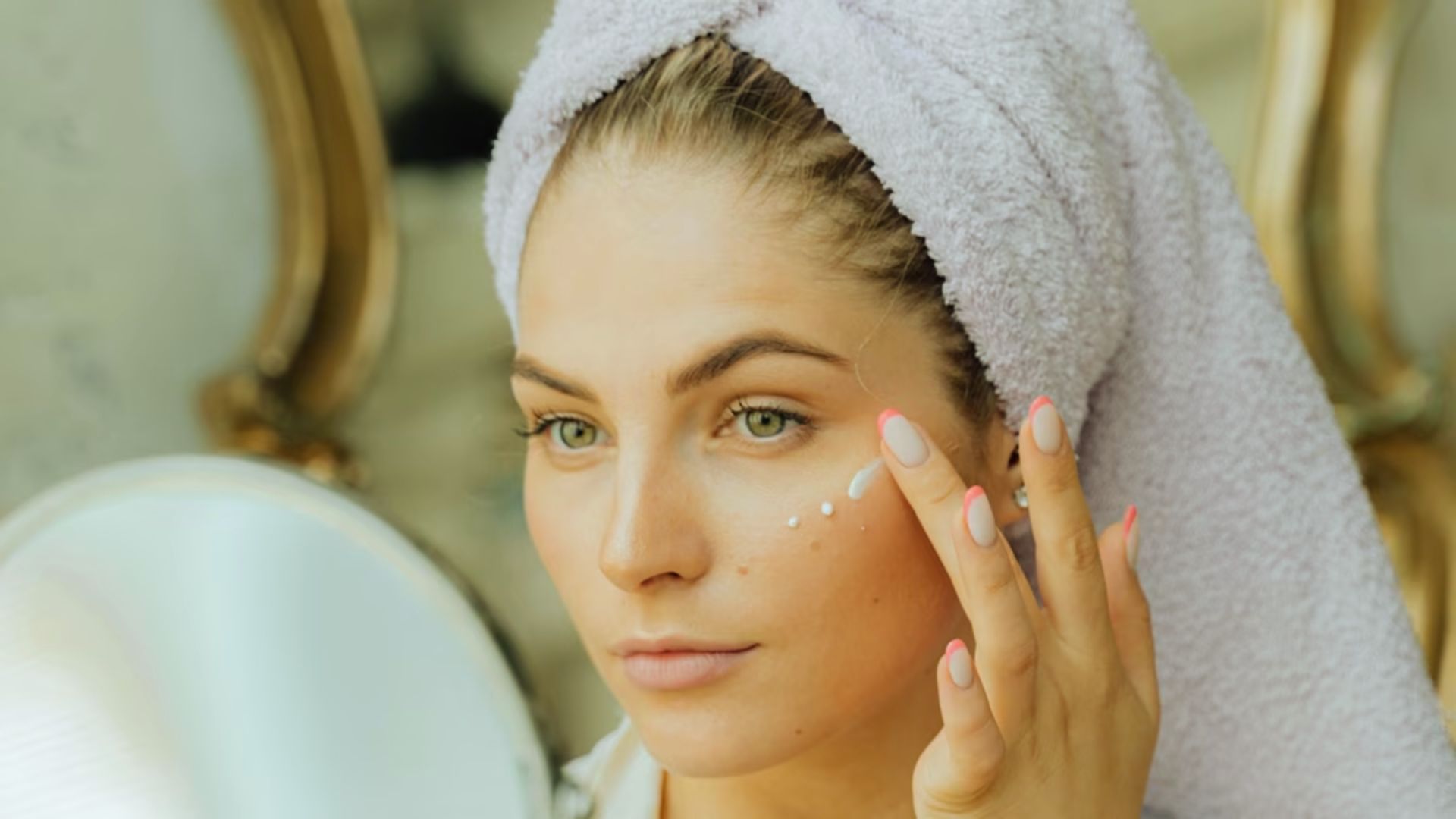 A woman wrapped in a towel applies cream to her face, focusing on skincare in a bright, serene bathroom setting.n