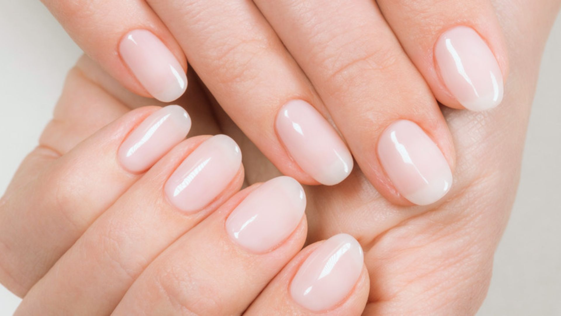 Close-up of hands with manicured nails in a soft, pale pink color. The nails are shiny and oval-shaped, conveying elegance and self-care.