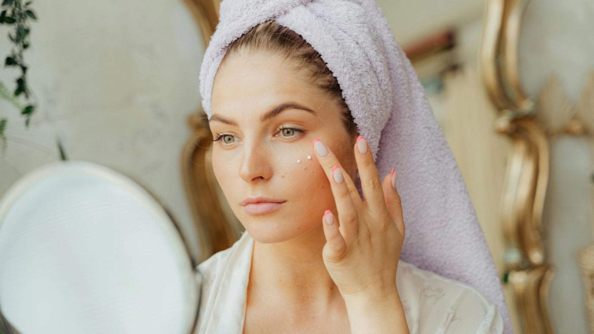 A woman wrapped in a towel examines her face in the mirror, focusing on her reflection.