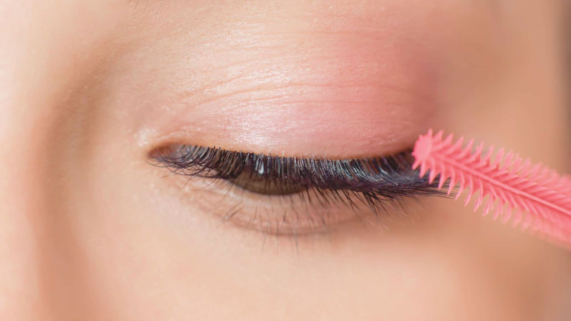 A woman applies mascara with a pink brush, focusing on enhancing her eyelashes.