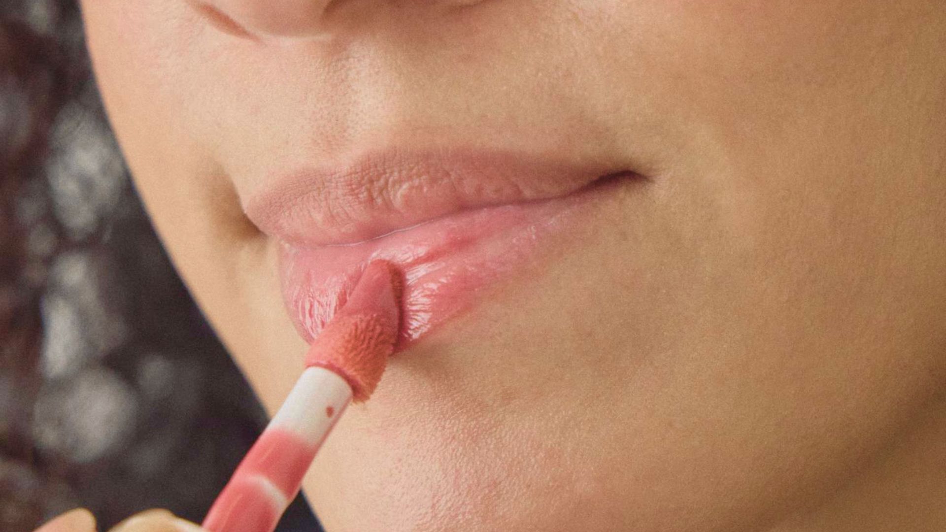 A woman applies lip gloss while brushing her lips with a pink toothbrush.