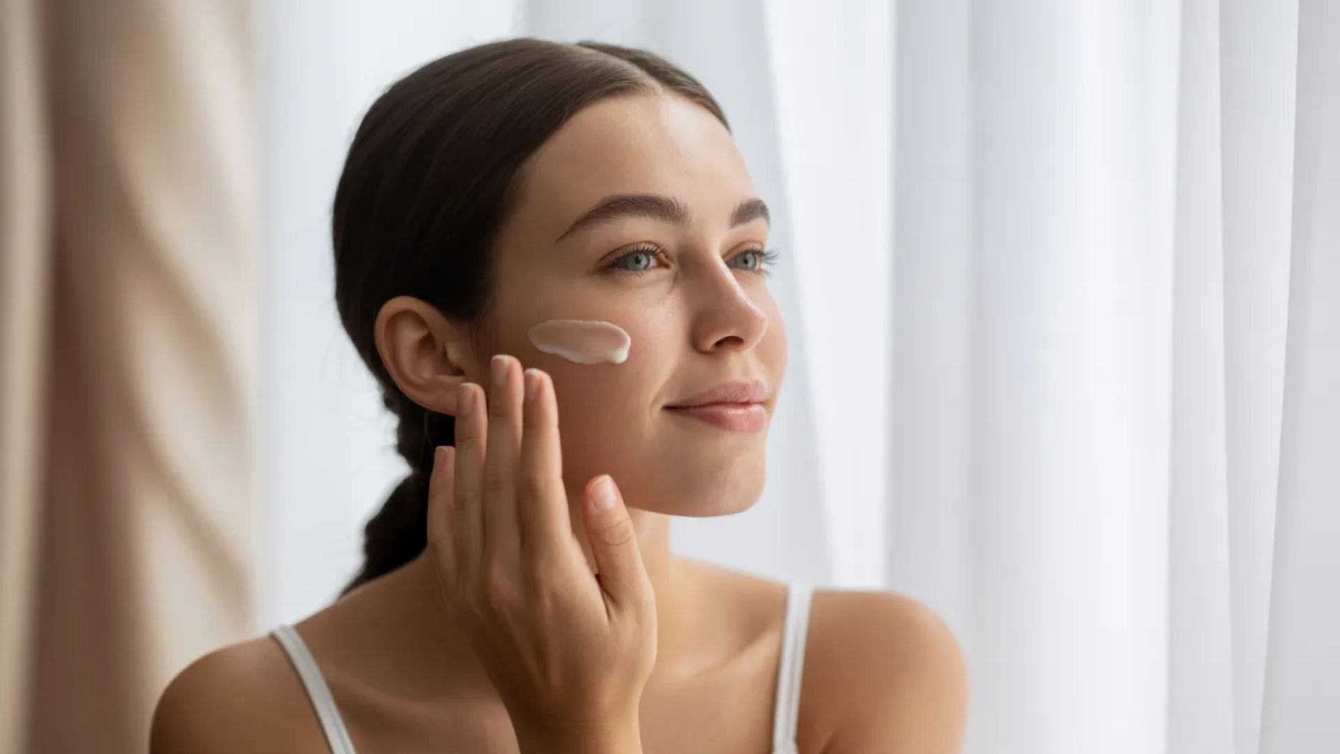 A woman is seen applying cream to her face, emphasizing her skincare routine in a bright, inviting environment.