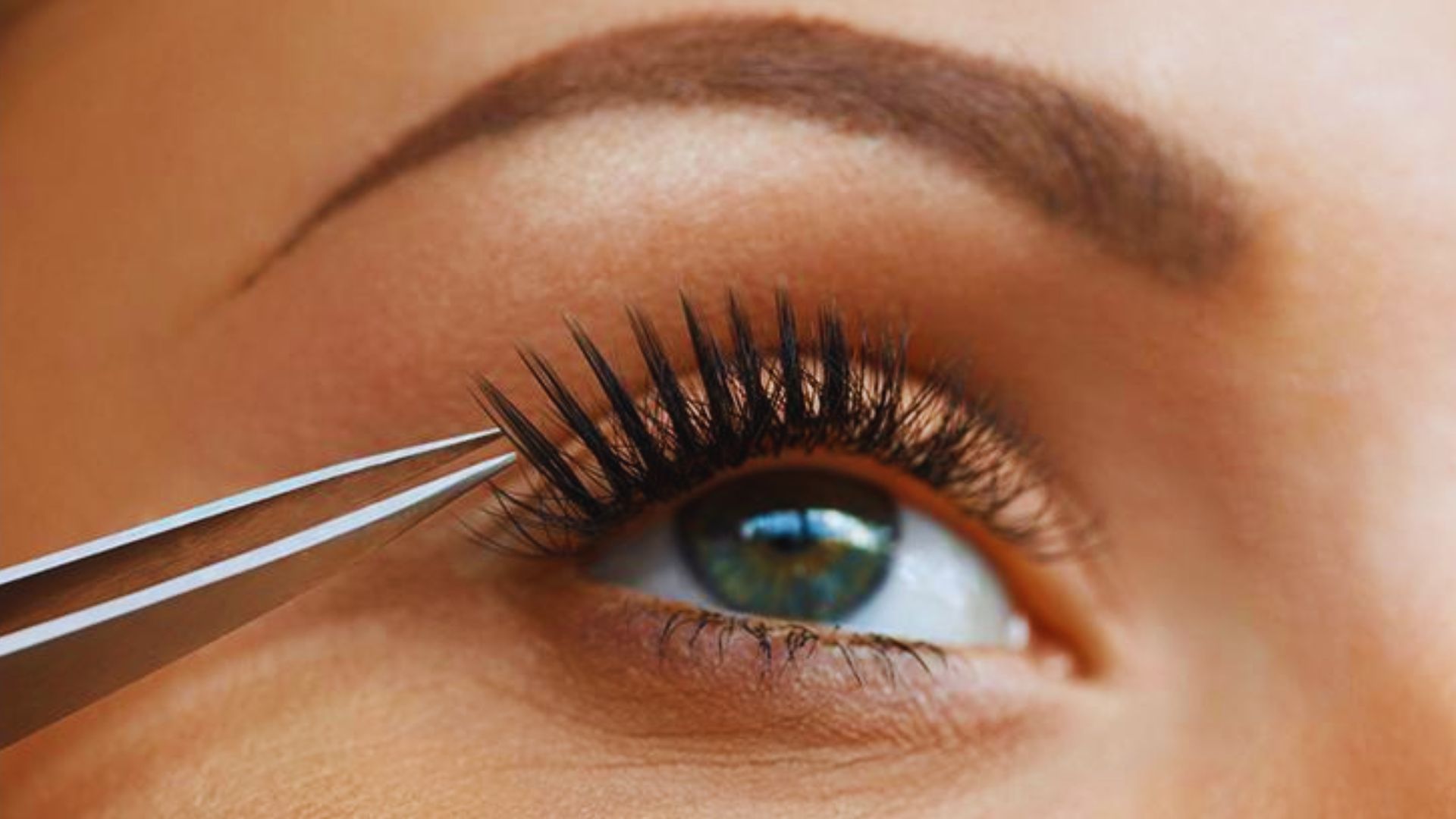 A woman with long eyelashes is seated while a beautician carefully trims her eyelashes.