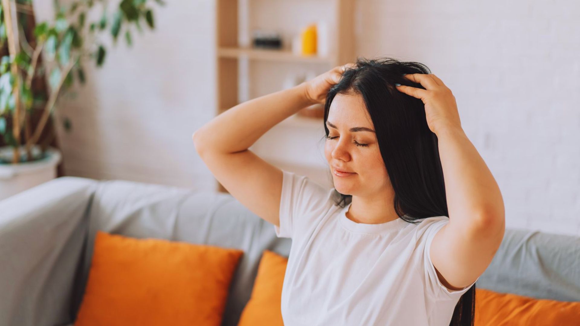 A woman sits on a couch, holding her hair with a thoughtful expression on her face.