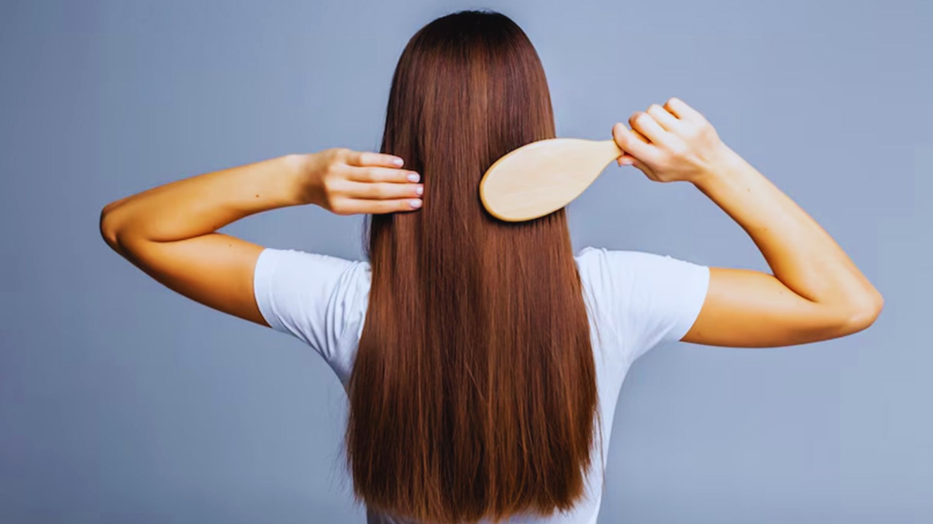 A woman with long hair is gently combing her hair, focusing on her reflection in a mirror.