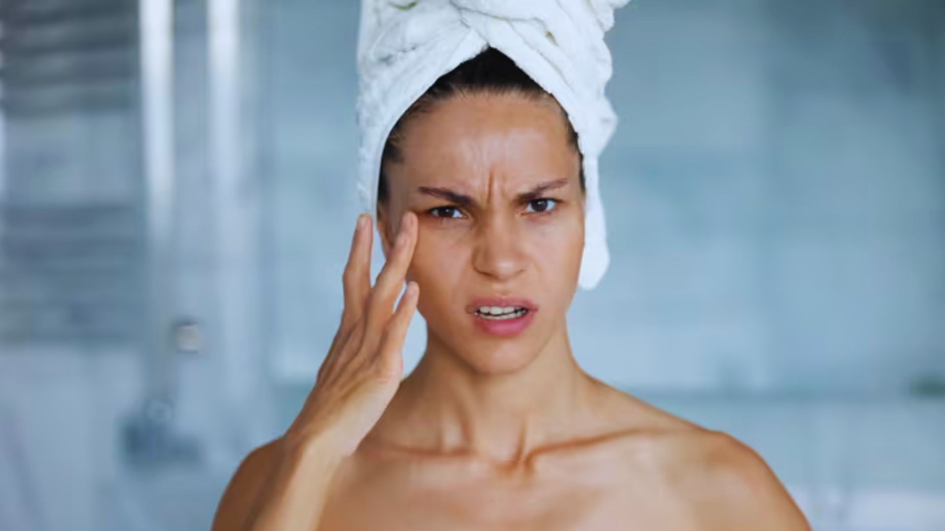 A woman wrapped in a towel gently touches her face, appearing relaxed and contemplative.