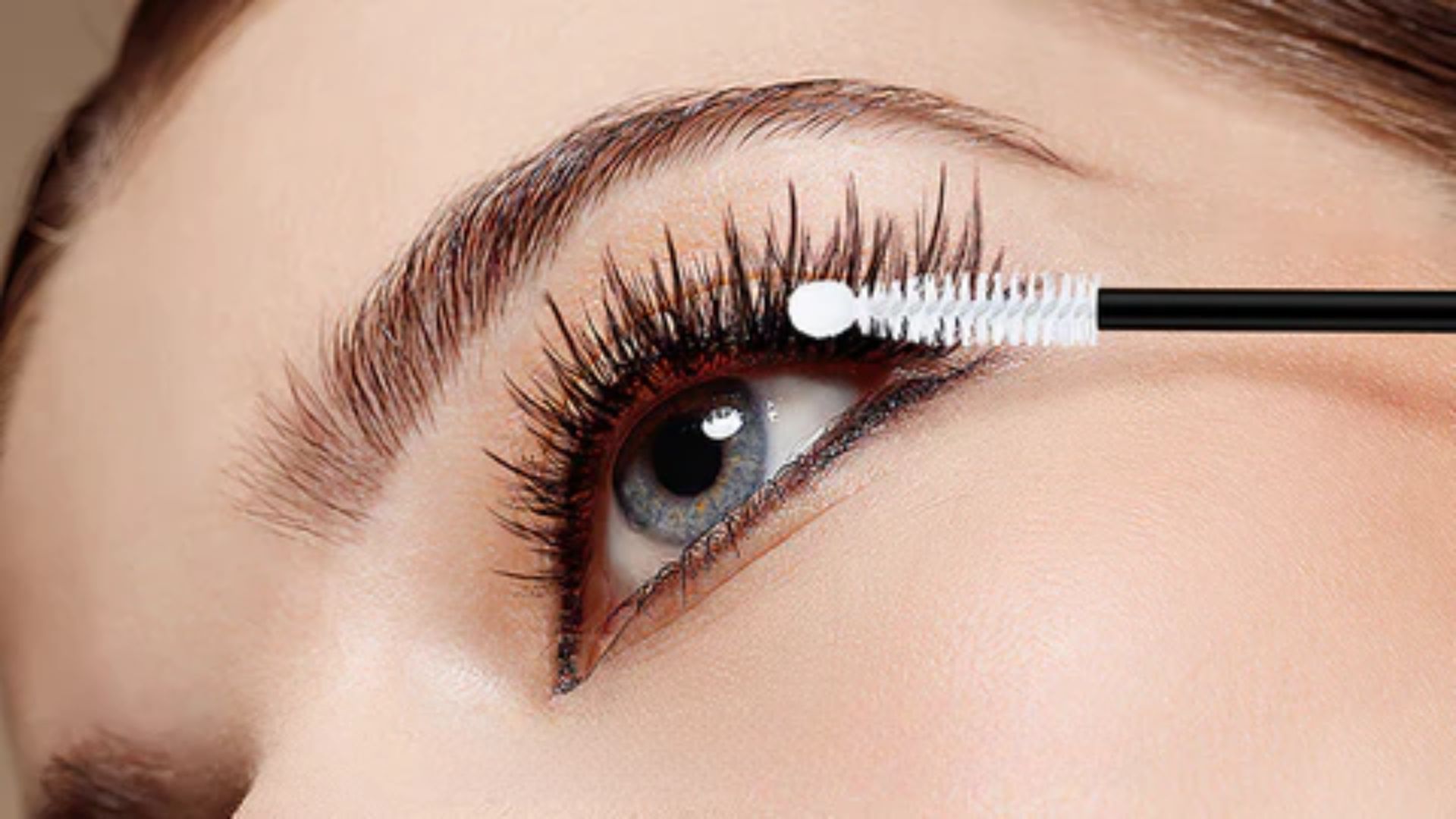 A woman applies mascara to her eyelashes using a mascara brush, focusing intently on her makeup application.
