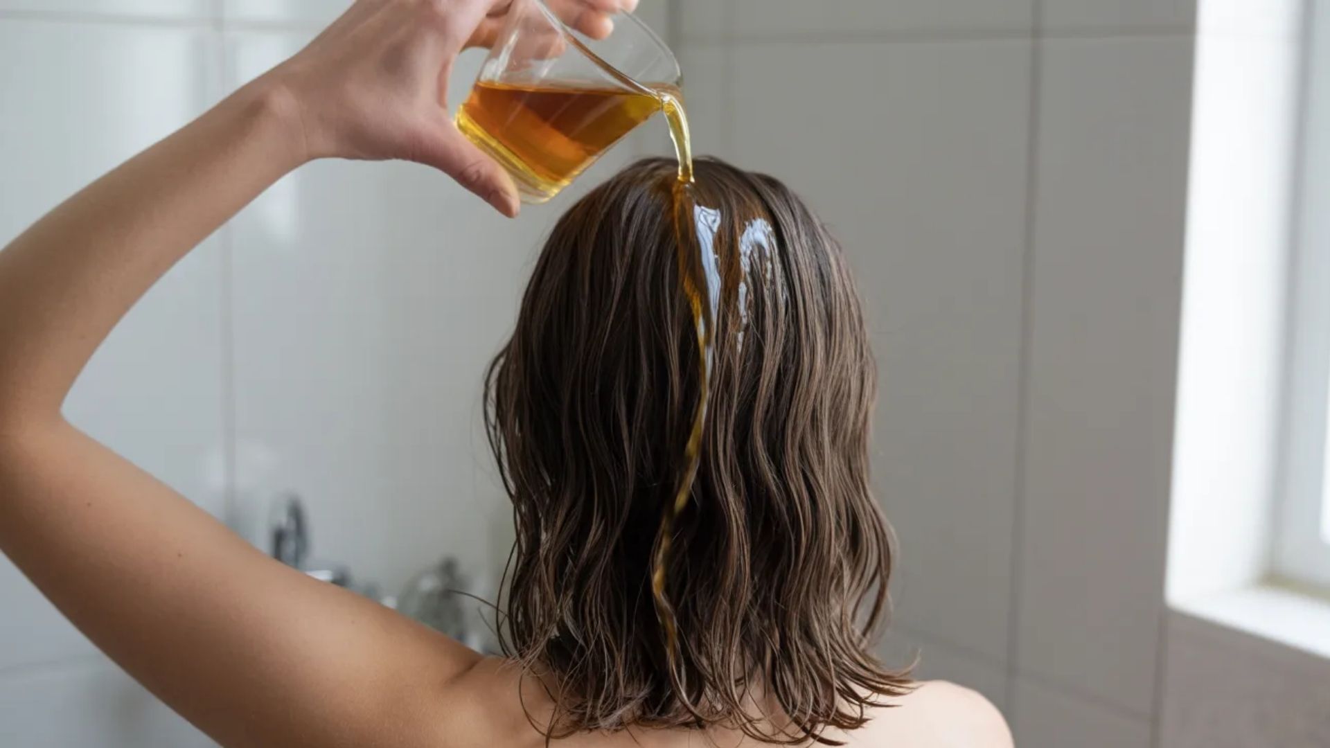 A woman washes her hair with a glass of apple cider vinegar rinse, promoting shine and scalp health naturally.