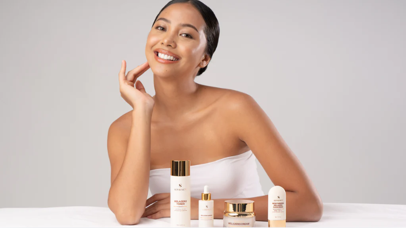 A smiling woman holds her skincare products, pondering whether to apply toner or serum first.