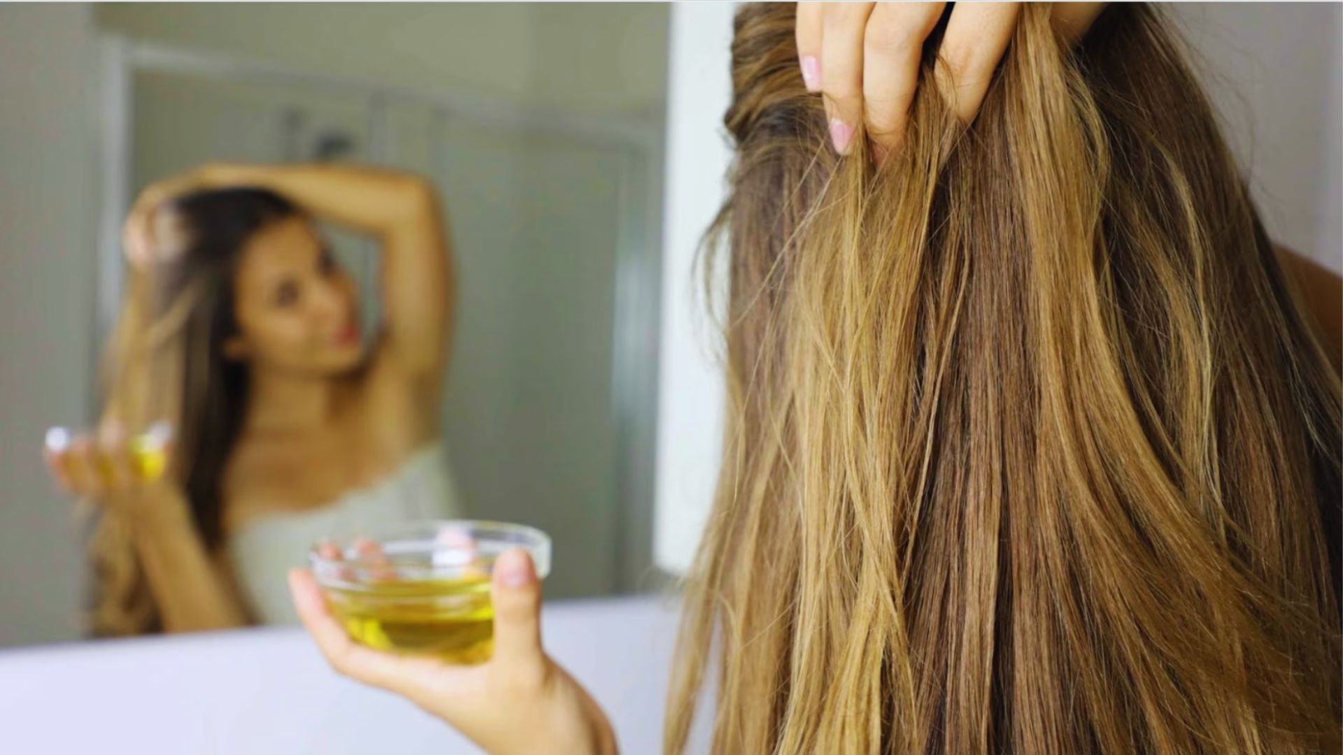 A woman combs her hair while applying castor oil from a bowl, focusing on hair care and nourishment.