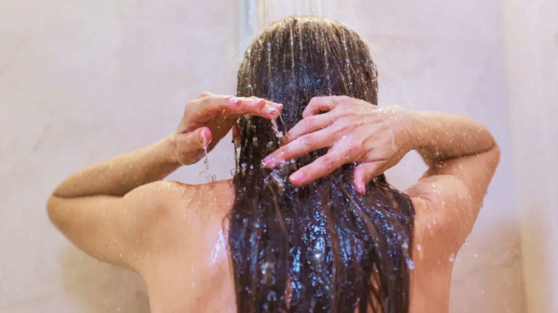 A woman washes her hair in the shower, surrounded by water and steam.