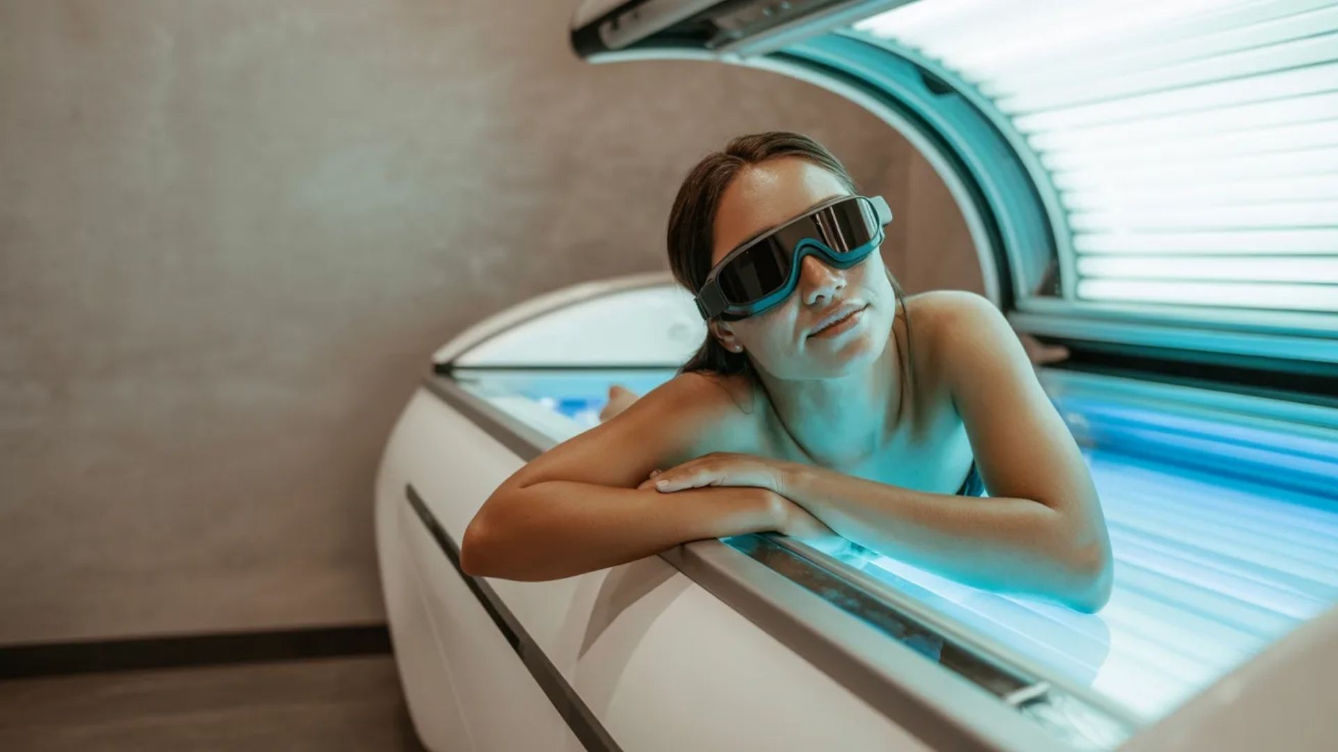 A young woman in sunglasses relaxes in a tanning bed, illuminated by soft blue UV light, wearing protective goggles.