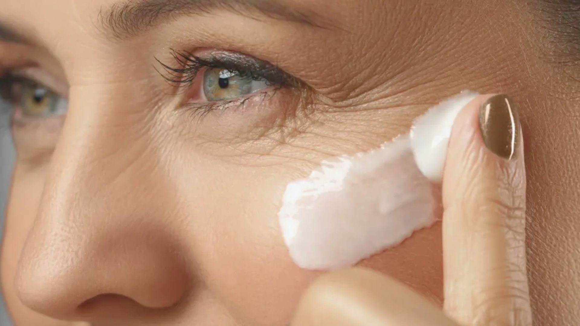 A woman gently applies cream to her face, focusing on skincare in a well-lit bathroom setting.