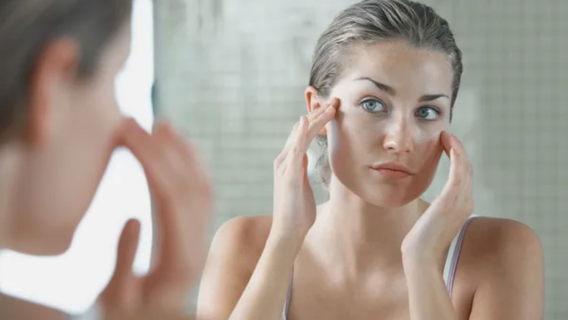 A woman applies skin care products while gazing into a mirror, focusing on her reflection and skincare routine.