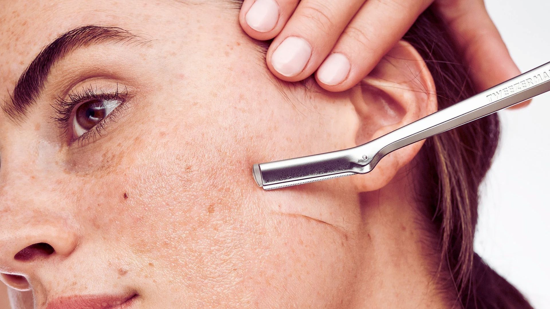 A woman carefully shaves her face with a razor, focusing on her grooming routine in a well-lit bathroom.