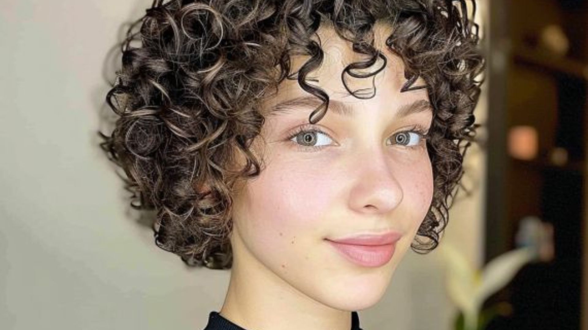 A young woman with curly hair gazes directly at the camera, smiling softly.