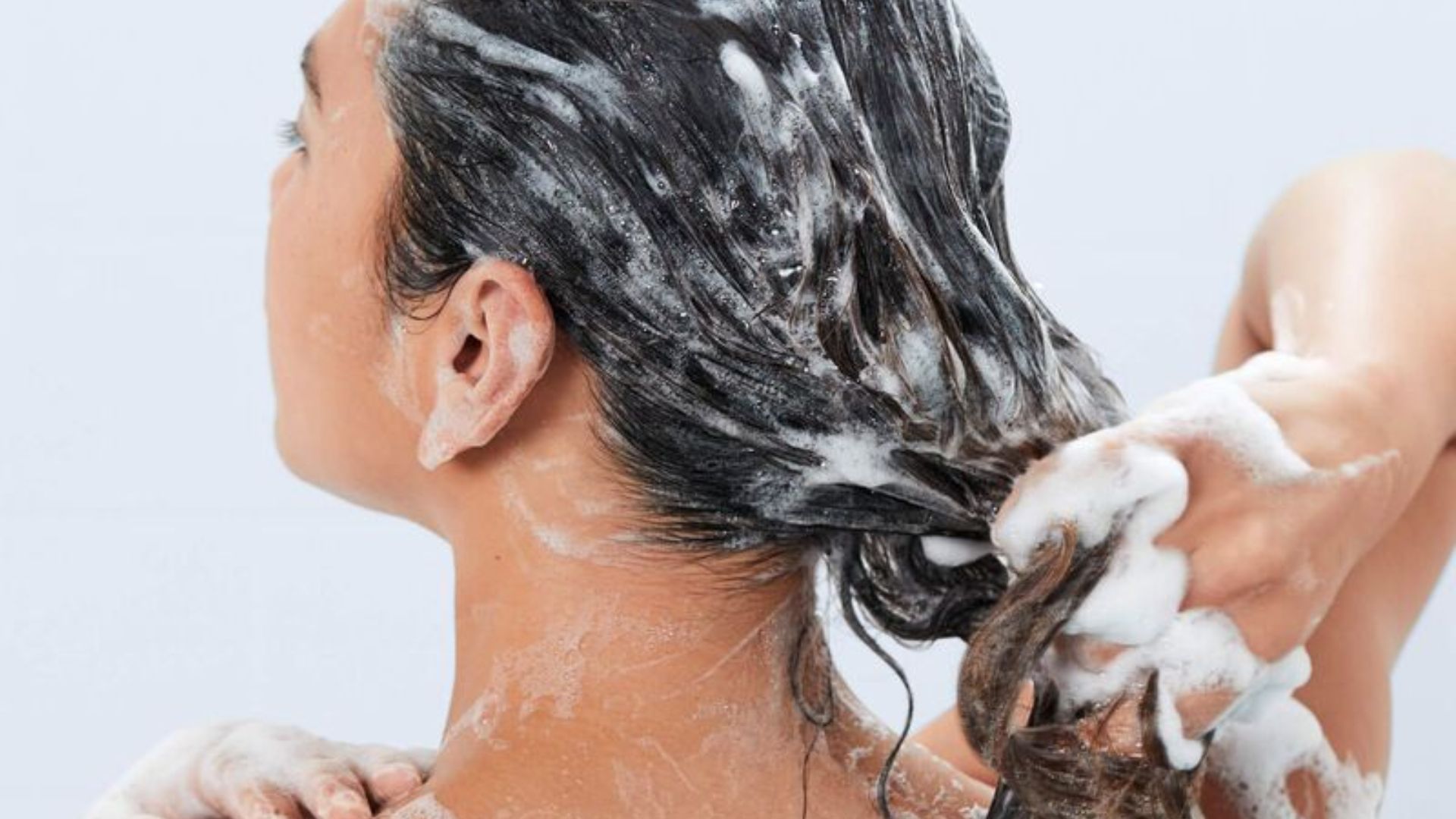 A woman washes her hair with soap, lathering it thoroughly in a bright bathroom setting.
