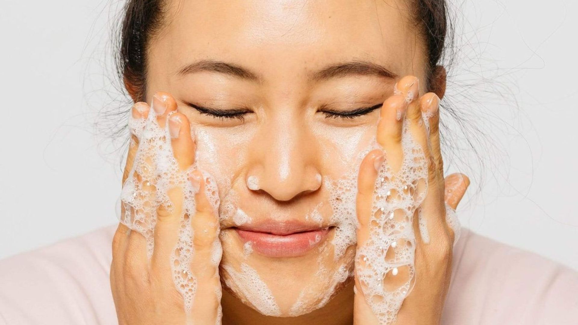 A woman is washing her face with soap, lathering her skin gently at a bathroom sink.