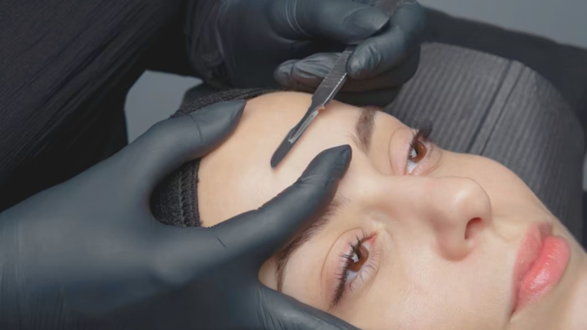 A woman sits in a salon chair while a beautician shapes her eyebrows with precision.