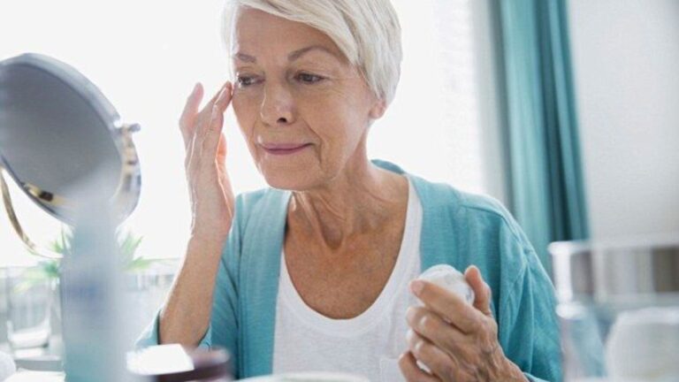 An older woman applies cream while gazing at her reflection in the mirror, focusing on her skincare routine.