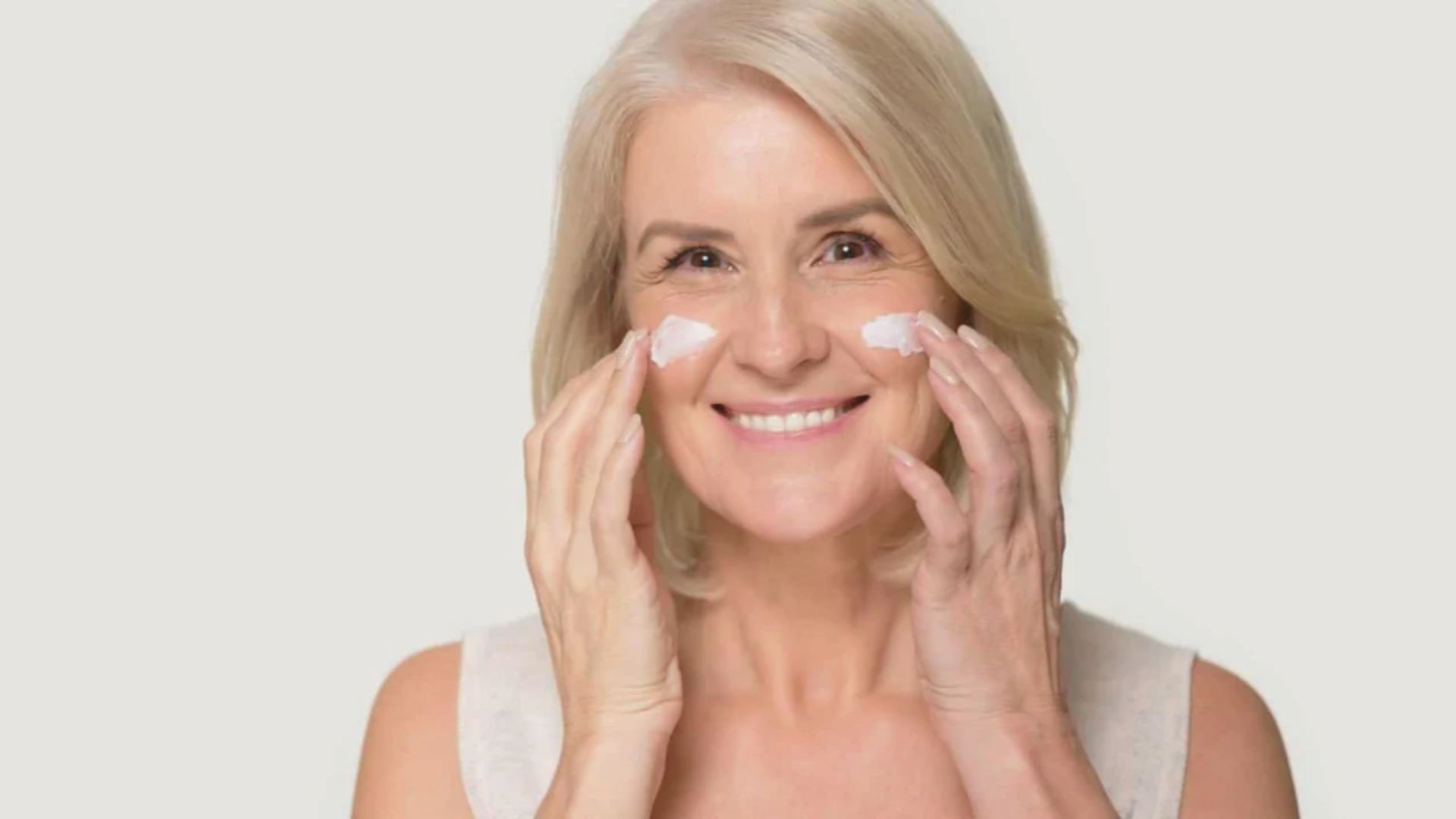A woman smiles while applying facial cream, showcasing a moment of self-care and skincare routine.