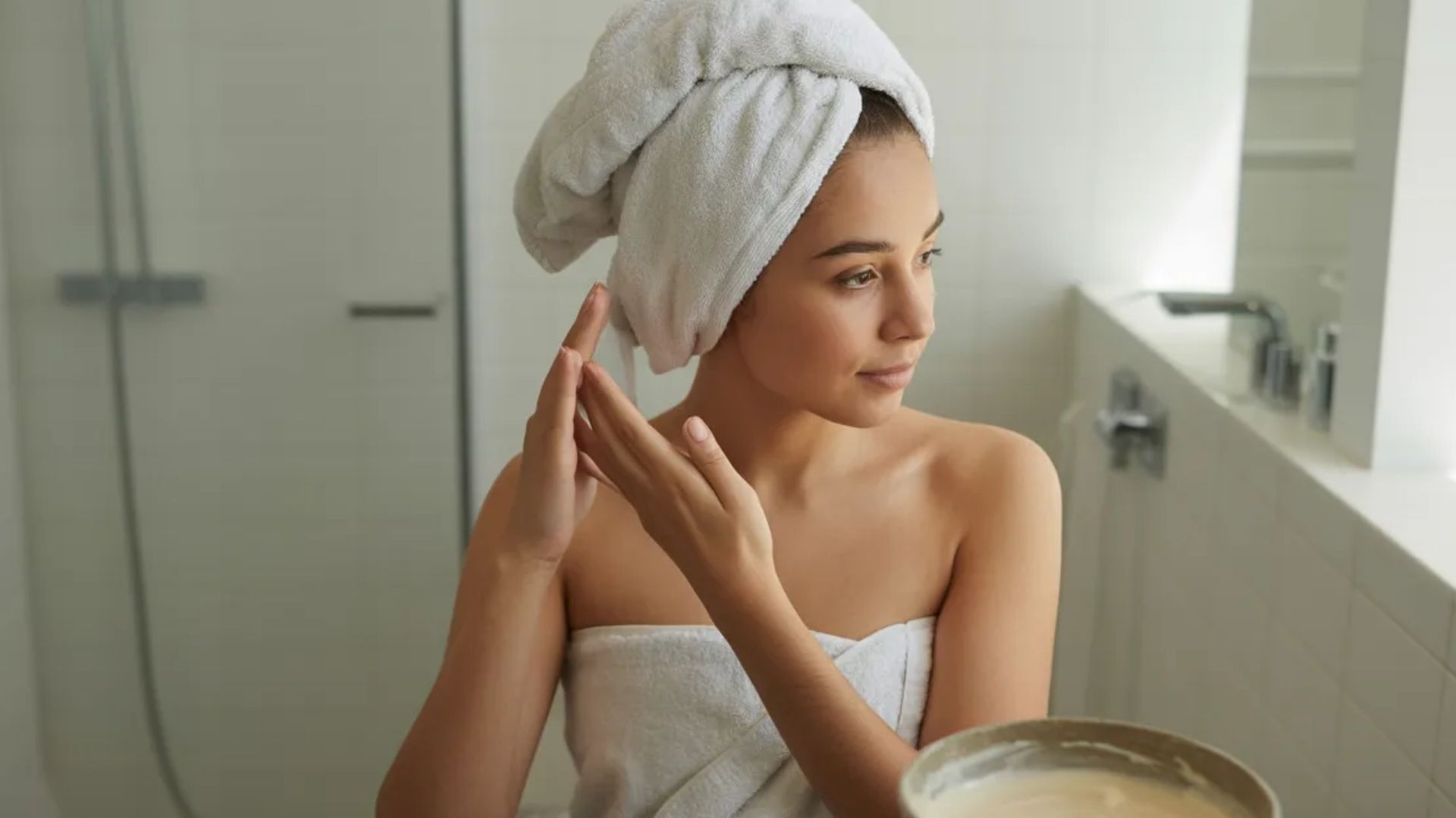A woman in a towel gently washes her face at a sink, emphasizing her skincare regimen in a bright bathroom.