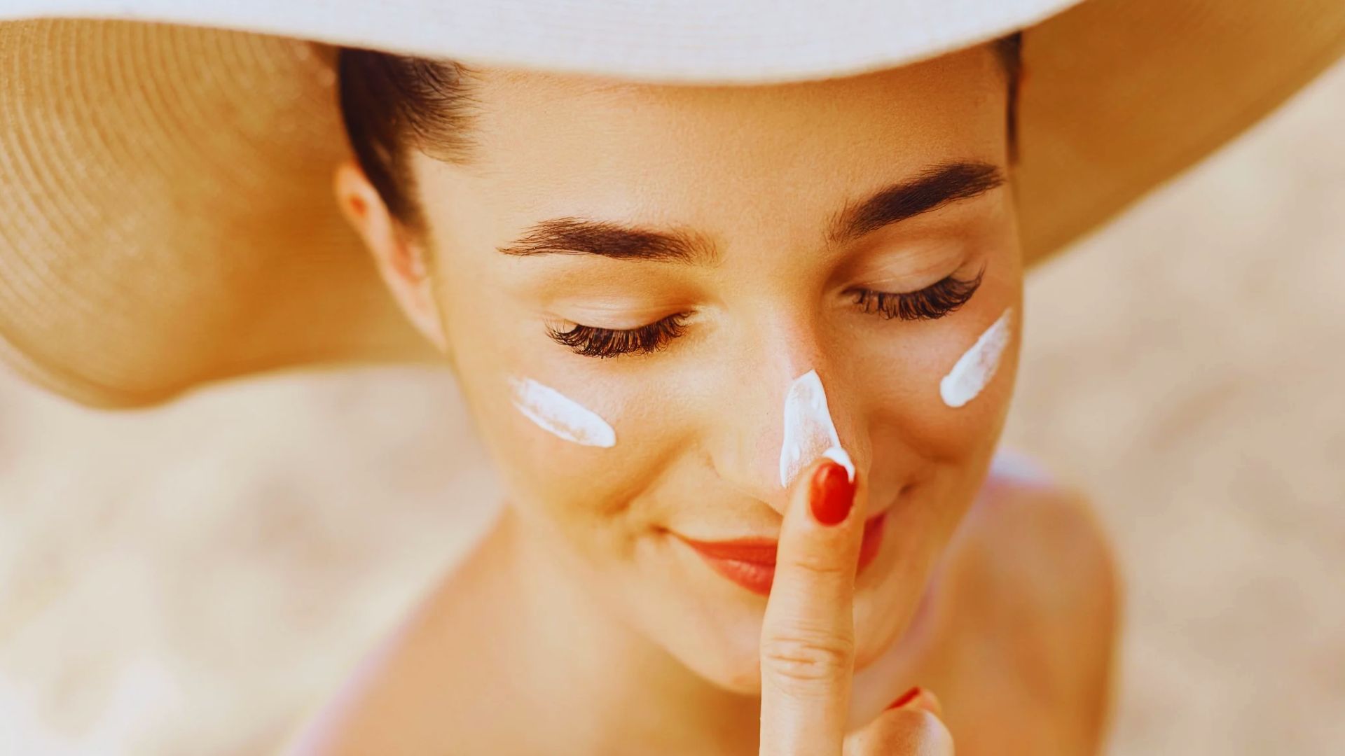 A woman wearing a hat applies sunscreen to her face, preparing for sun exposure.