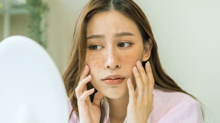 A woman gazes at her reflection in a mirror, examining her face with a thoughtful expression.
