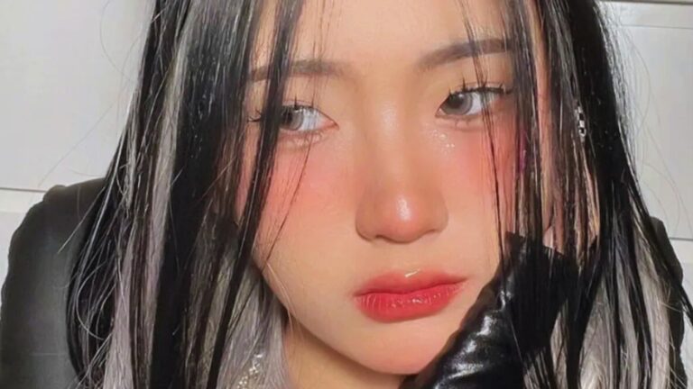 Young woman with long, straight black hair and silver streaks gazes to the side. Her expression is contemplative, with a subtle blush and glossy lips.