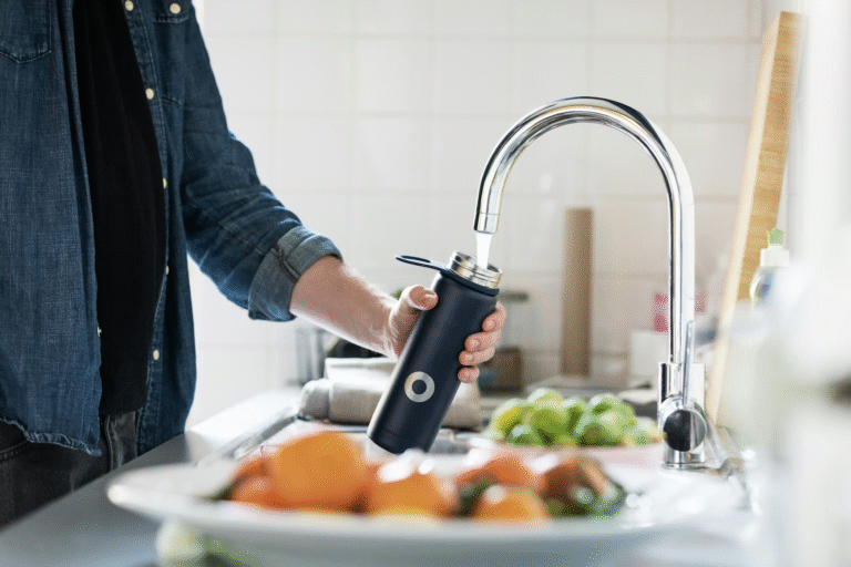 A person in a denim shirt fills a navy reusable water bottle at a kitchen sink. Fresh oranges sit in the foreground, with a bright, welcoming tone.