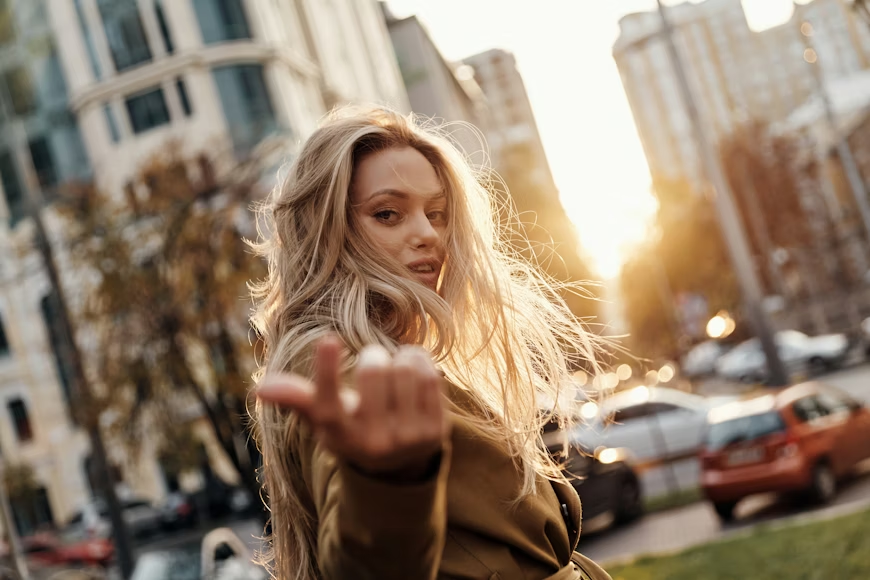 Woman with long blonde hair in a trench coat gestures to follow, with a cityscape and golden sunset behind her, conveying a sense of invitation and adventure.