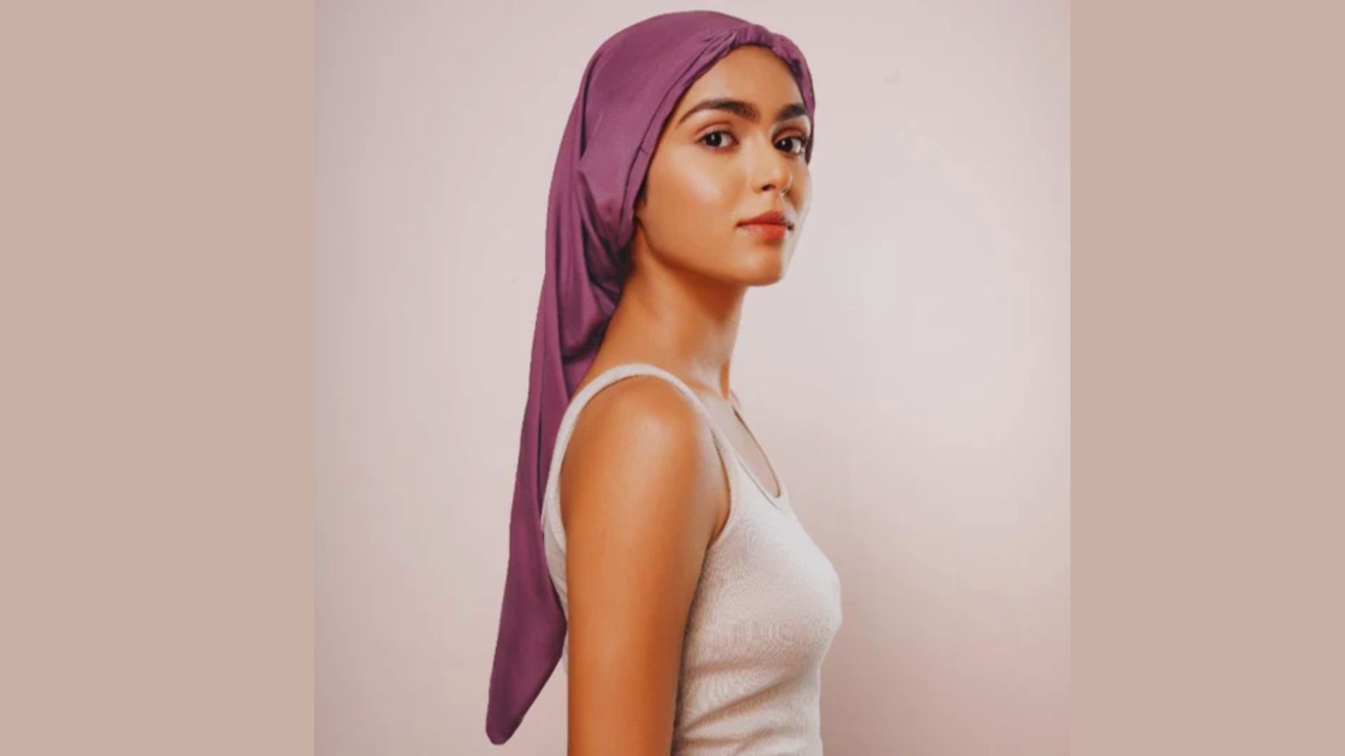 A woman with a purple headscarf, smiling softly, stands against a neutral background.