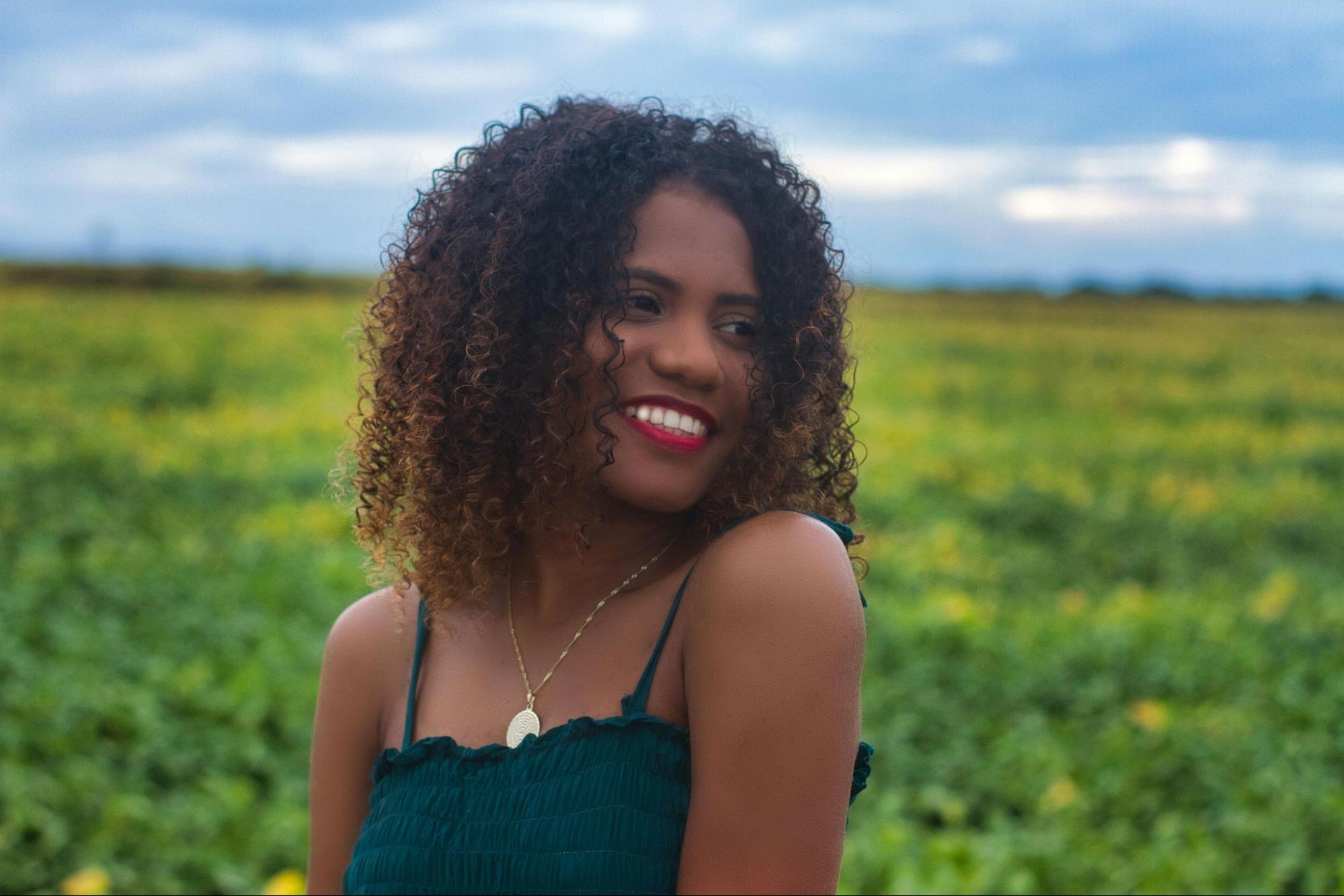 A woman with curly hair and a bright smile stands in a field of green plants under a cloudy sky, conveying a joyful and serene mood.