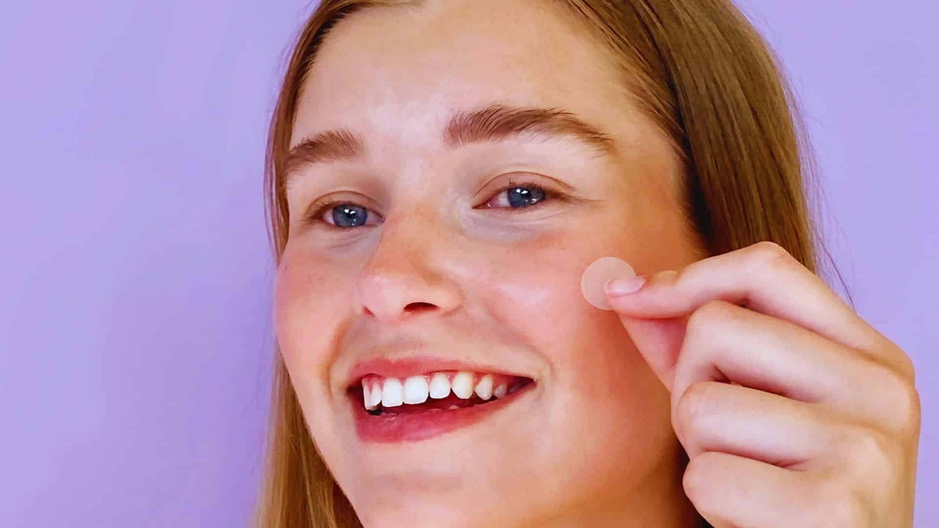 A woman smiles as she applies cream to her face, showcasing a moment of self-care and beauty routine.