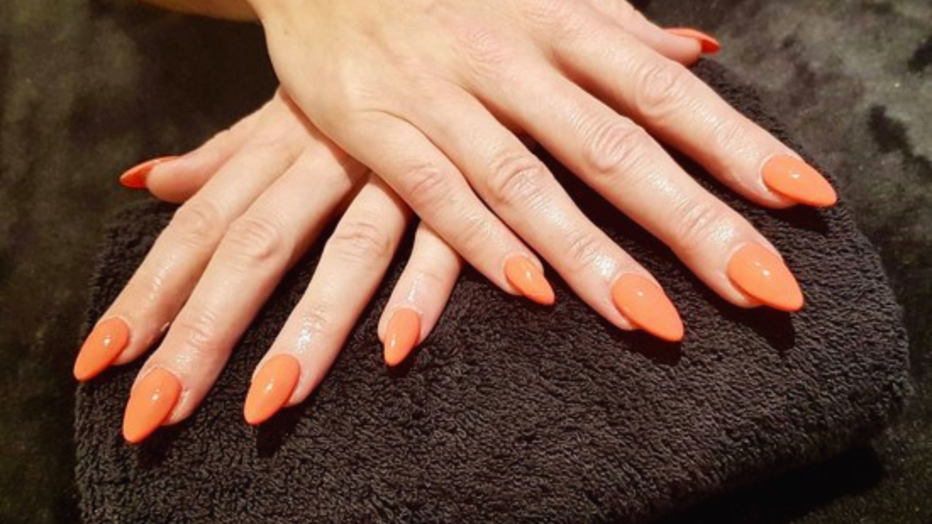 Bright orange nails resting on a black towel, showcasing a vibrant manicure against a dark background.