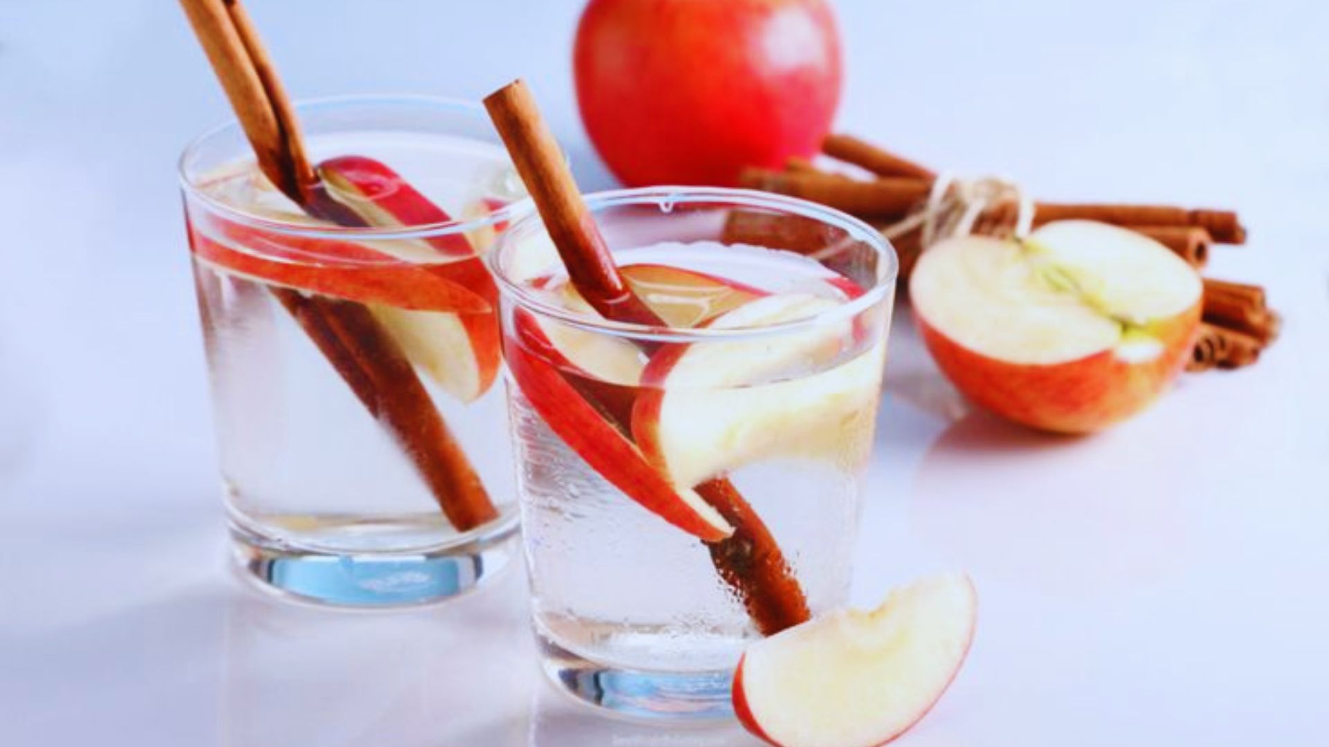 Two glasses of water beside fresh apples and cinnamon sticks on a wooden table.