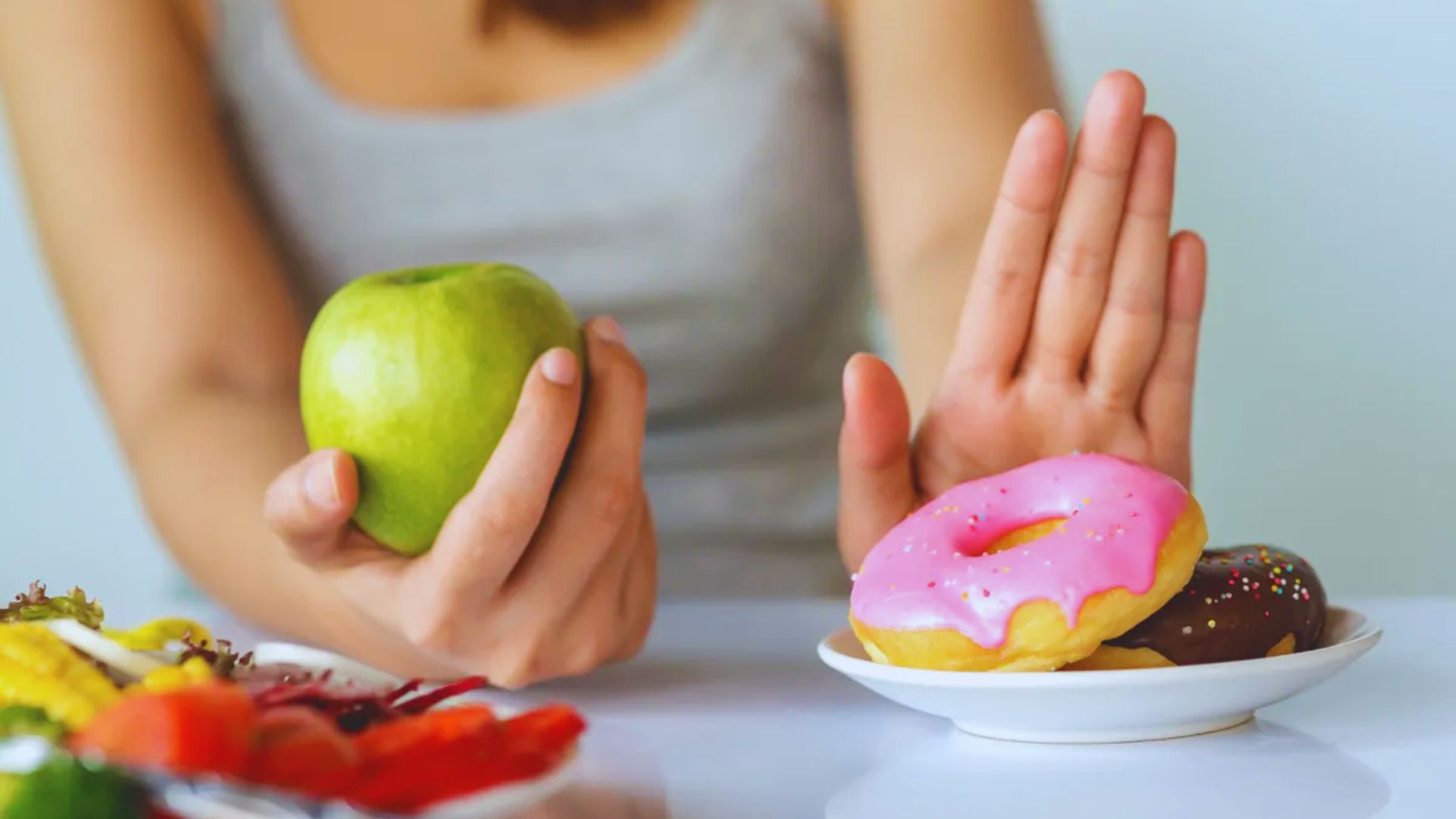 A woman holds an apple and a donut, standing in front of a plate filled with various foods.