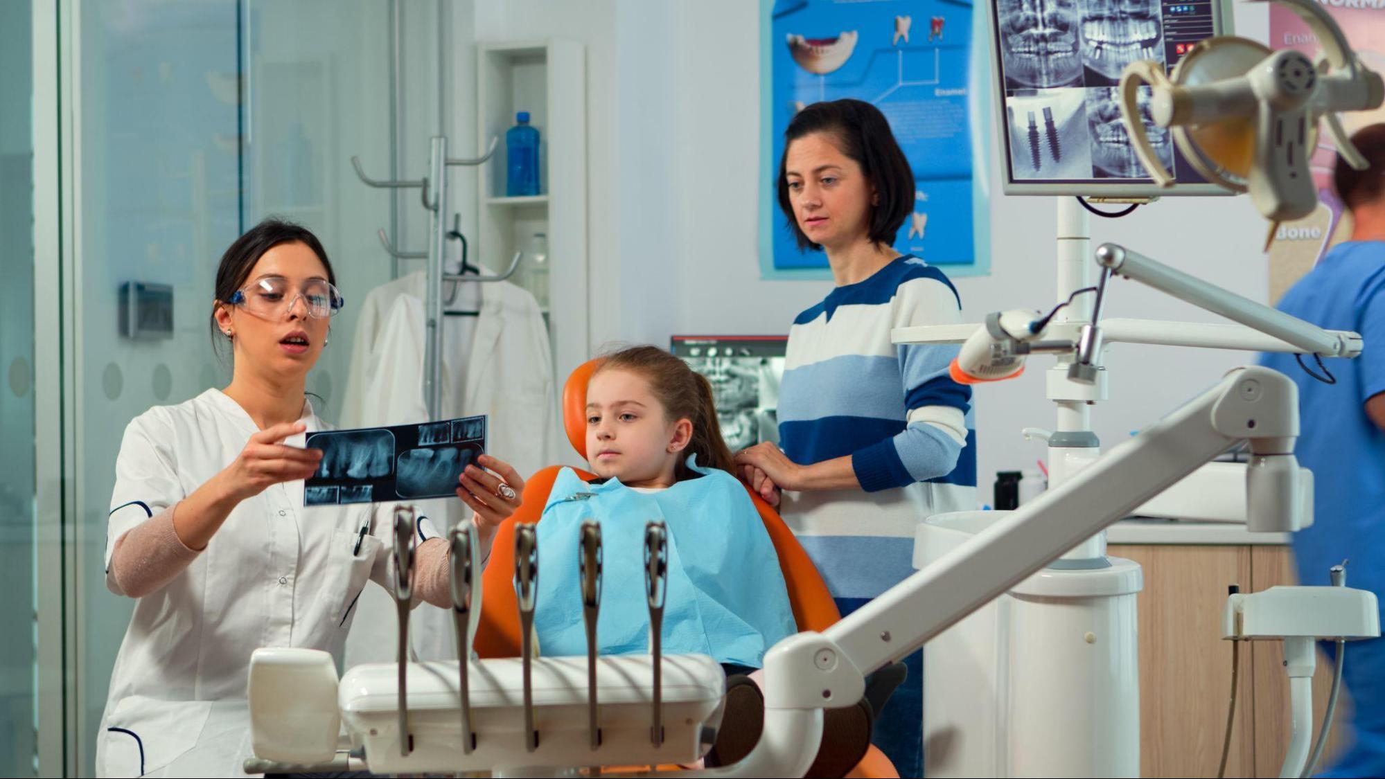 A dentist explains an X-ray to a young girl in a dental chair, while her concerned mother listens. The room is bright, with dental equipment visible.