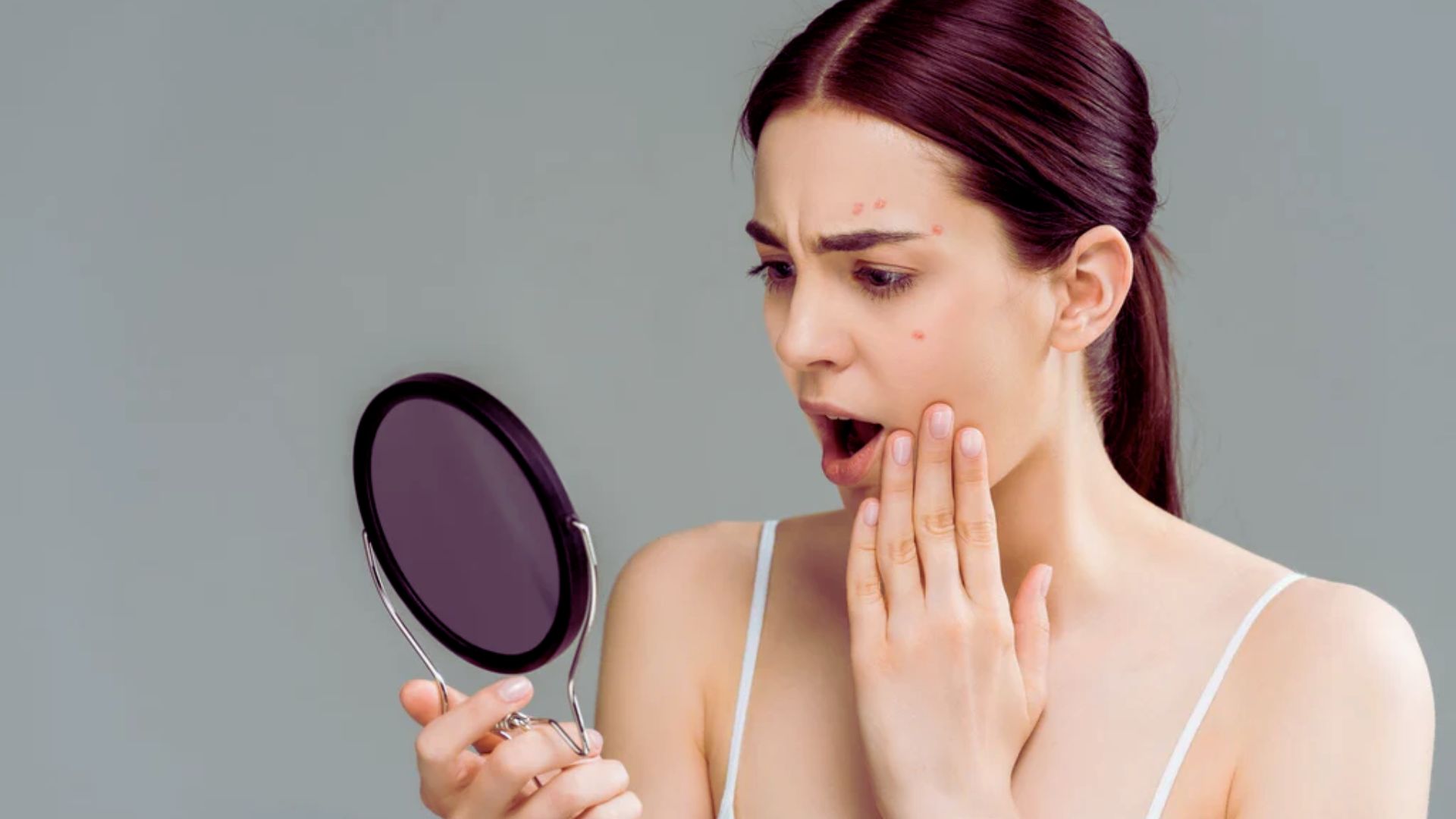 A woman gazes at her reflection in a mirror, examining her face with a thoughtful expression.