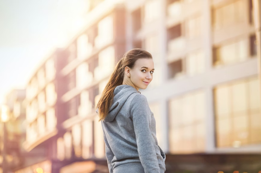 A young woman in a gray hoodie smiles while turning back, standing in front of blurred modern buildings at sunset, conveying a sense of calm and contentment.
