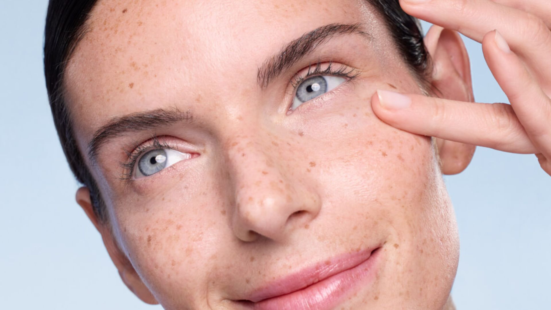 A woman with freckles gently touches her eye, showcasing her natural beauty and delicate features.
