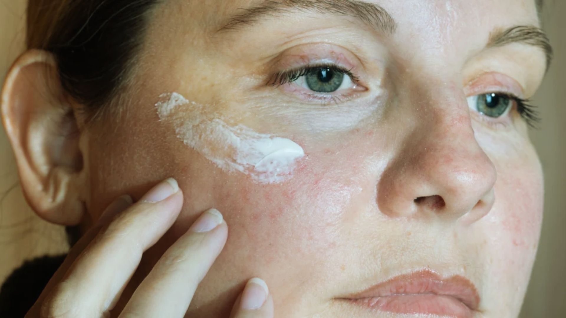 A woman applies white cream to her face, focusing on skincare in a bright, well-lit environment.