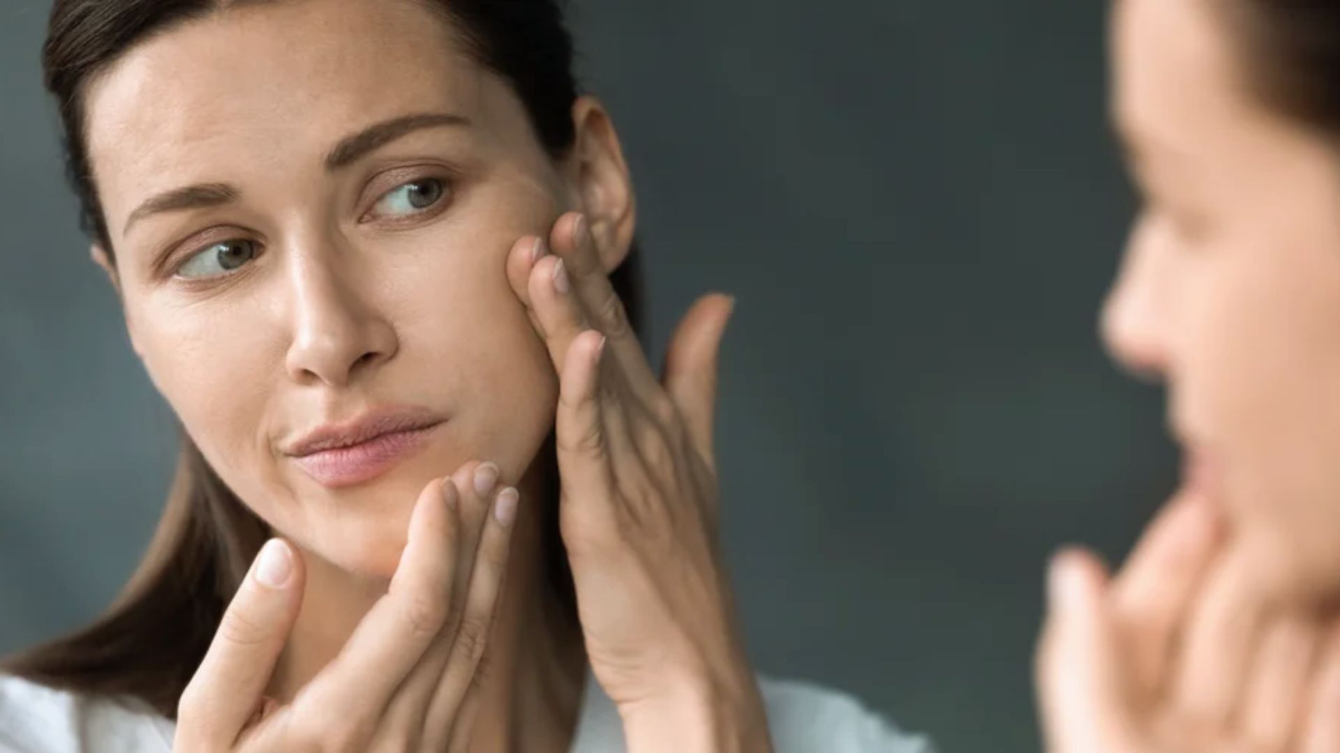 A woman gazes at her reflection in a mirror, examining her face with a thoughtful expression.