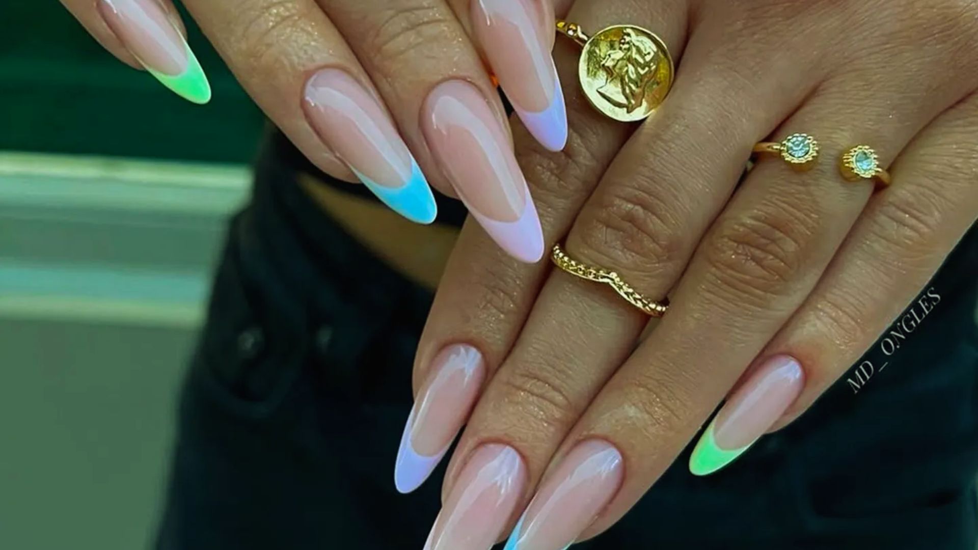 A woman displays her long nails painted in vibrant blue and green colors, showcasing a stylish manicure.