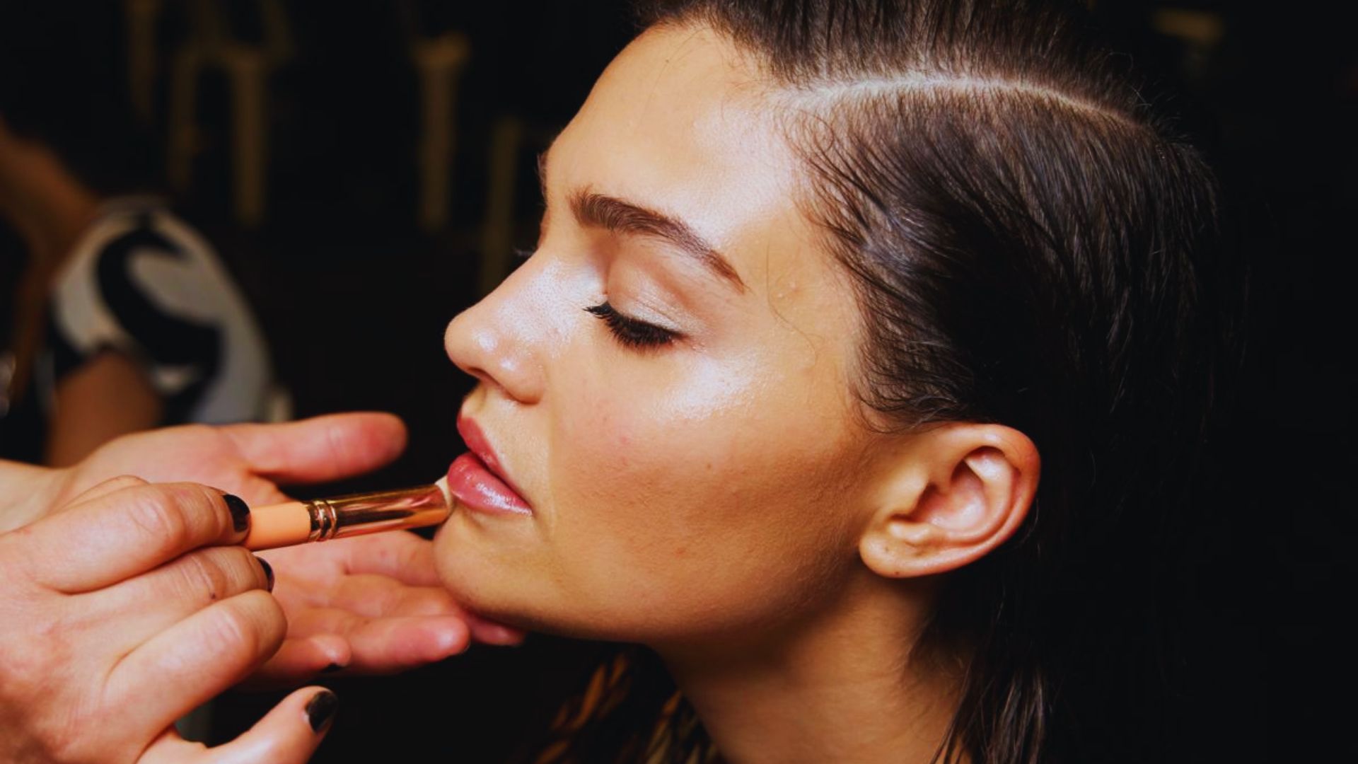 A woman receiving makeup application backstage at a fashion show, surrounded by stylists and fashion elements.